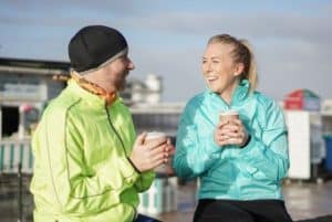 A young mand and young woman sitting outside wearing exercise clothes, smiling at each other and holding a hot drink.