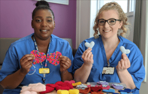 Two medical professionals wearing blue scrubs, sat next to each other smiling at the camera and holding heart shaped knitted items.