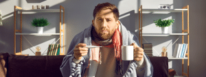 A man sat on the sofa looking unwell. He is wrapped in a blanket and holding a thermometer and mug.