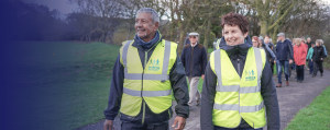 A group of people walking on a path through a park. A man and woman are at the front of the group, both wearing high viability jackets with the 'walking for health' logo. They are smiling.